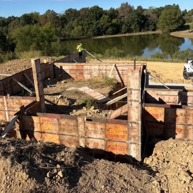 Steel wall forms set in a dig beside a pond, with a crew member on top